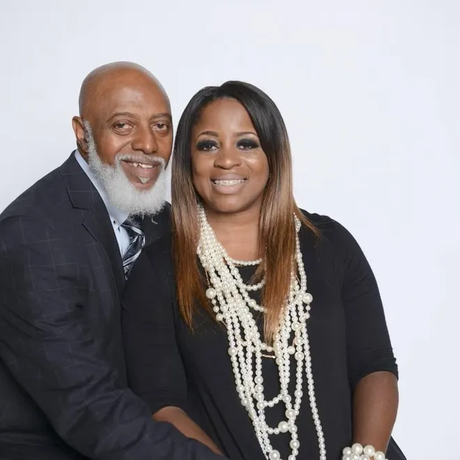Bishop Rodney Gamble Sr. smiling beside a woman in a black dress and layered pearl necklaces
