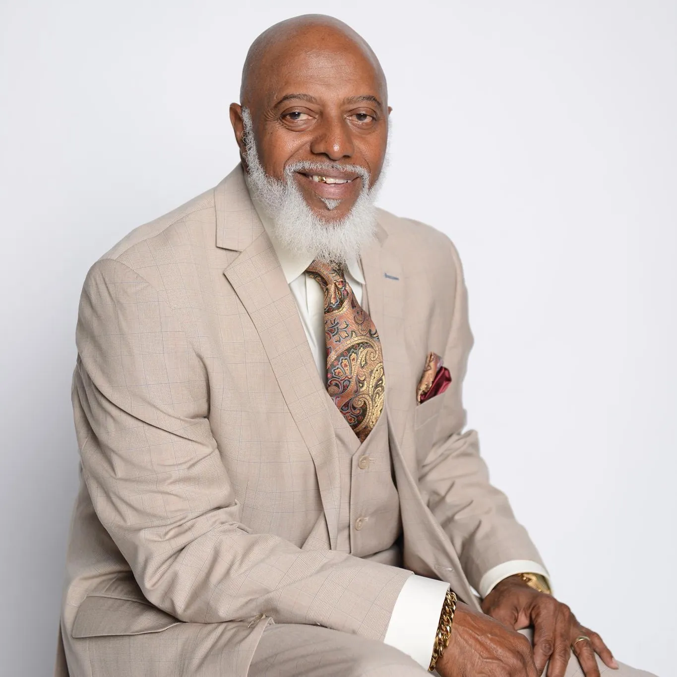 Bishop Rodney Gamble Sr., smiling, seated in a tan three-piece suit with a paisley tie and gold accessories
